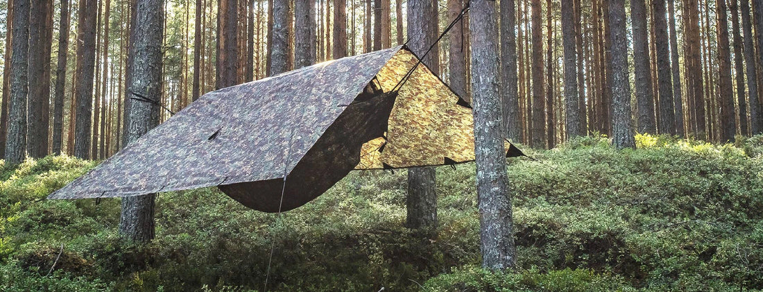 Camo camping tarp set up among trees in a serene forest, ideal for bushcraft and wild camping adventures.