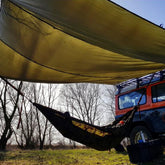 Bushmen Thermo Tarp 4x4 Olive used as shelter over a hammock by a vehicle in a natural setting.