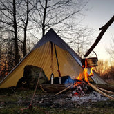 Camping setup with a pyramid tent, backpack, and a fire in a forest during sunset.