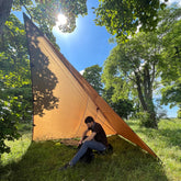 A person relaxing under a DD Tarp 4x4 shelter in a lush outdoor setting, showcasing its versatility for camping.