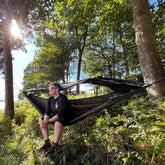 Man relaxing in a DD Frontline KING Size hammock in a forested setting, enjoying nature and comfort.
