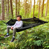 A child sitting in a DD Hammocks Junior Frontline Hammock, surrounded by lush green ferns in a forest.