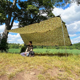 Person relaxing under DD Tarp XL in a natural setting, showcasing its ample shelter and camouflage design.