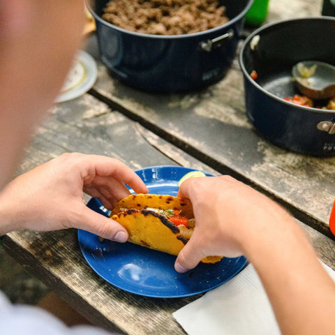 Person holding a taco on a blue plate with cooking pots in the background