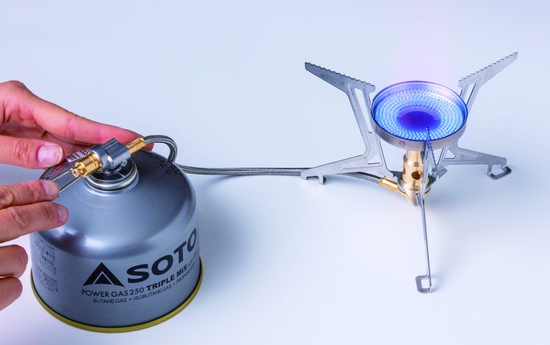 Hand holding a Soto gas canister to a camping stove on a white background